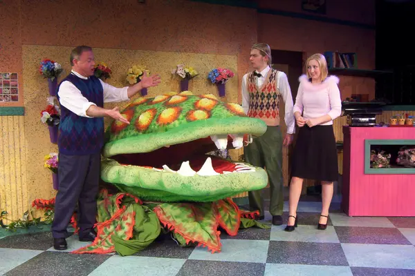 Stage play with actors and a large, colorful plant prop.