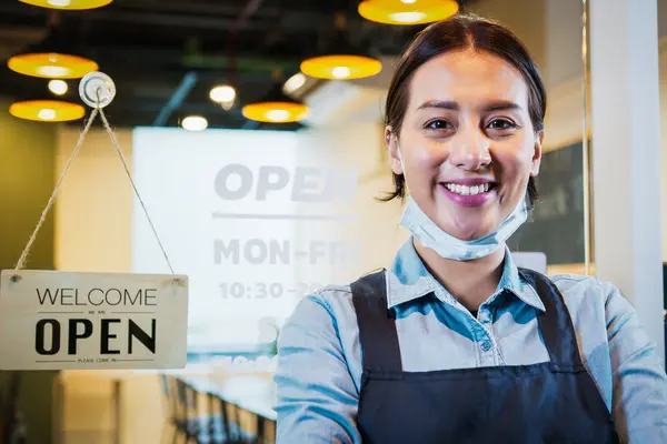 small business owner in front of open sign smiling with mask off of face