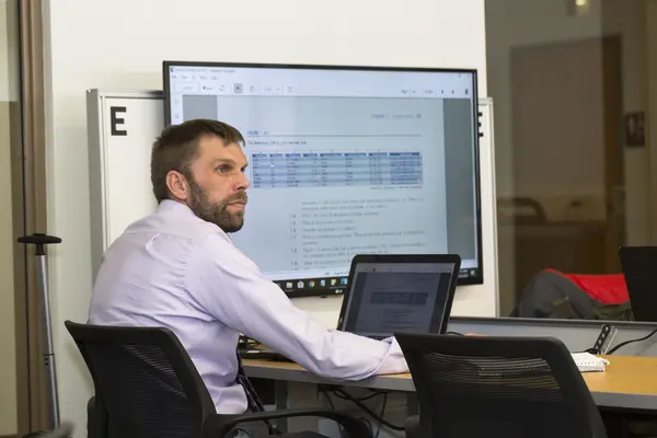 Male student in web development class, looking at laptop screen with data display in background.