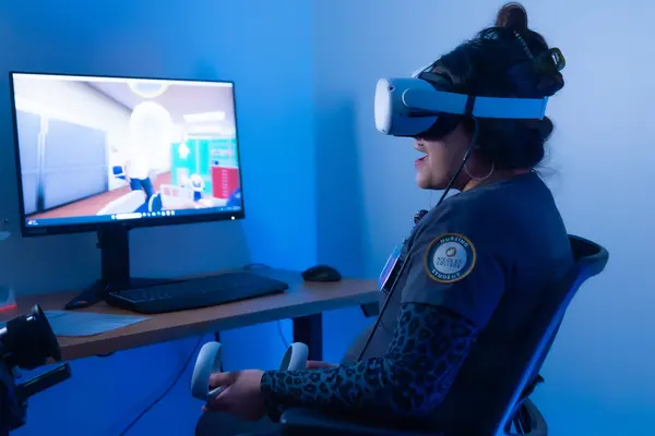 Person using VR headset and controllers, seated by a computer monitor.