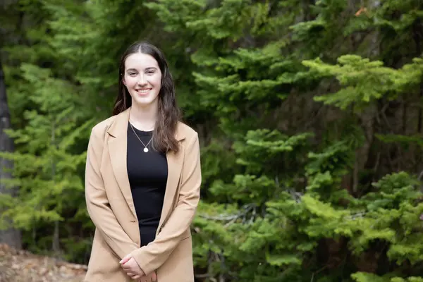 Woman smiling in front of lush green trees, wearing a beige coat and black top.