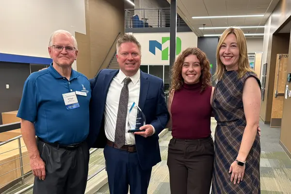Four people smiling in a well-lit hallway; one holds an award.