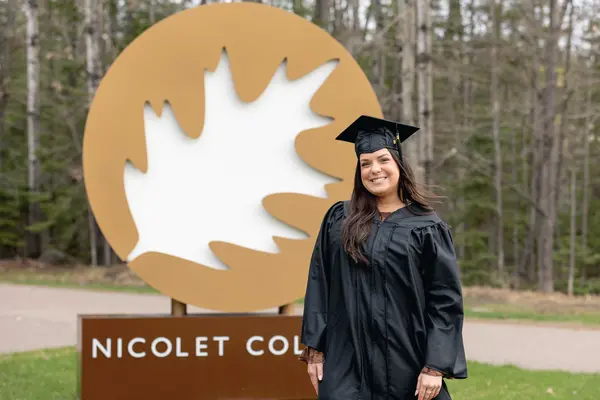 Woman with long brown hair smiling in a graduation cap and gown in front of a large Nicolet College sign.