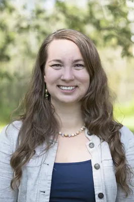Woman smiling outdoors in a light-colored jacket, with blurred trees in the background.