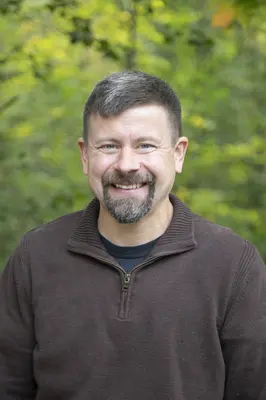 Smiling man in a brown sweater with a green outdoor background.