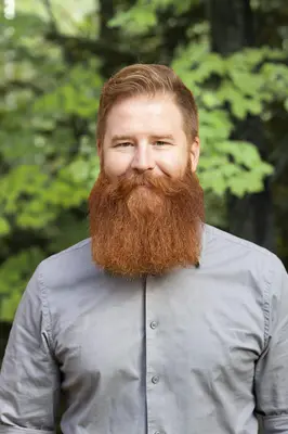 Man with a red hair and beard smiling in a forest setting.