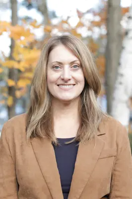 Smiling woman in a brown coat outdoors with autumn leaves.