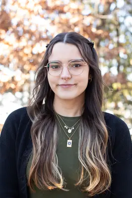 Young woman with glasses, wearing a green top and black cardigan, outdoors in autumn.