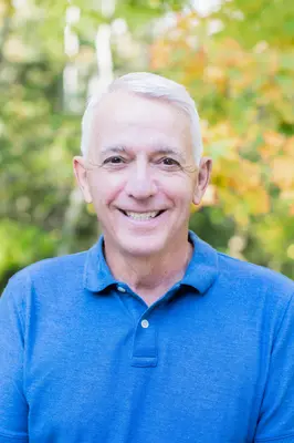 Smiling man in a blue shirt, outdoors with autumn leaves in the background.