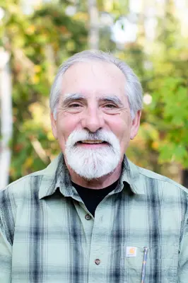 Smiling man with gray hair and beard, wearing a plaid shirt, outdoors.