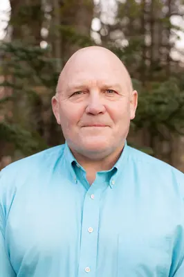 Bald man in blue shirt standing outside with trees in the background.