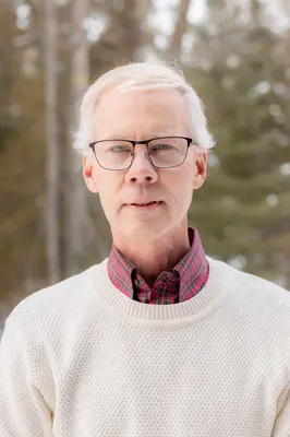 An older person with glasses in a white sweater, outdoors in a forest.