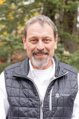 Man smiling outdoors, wearing a black vest and white shirt, trees in background.