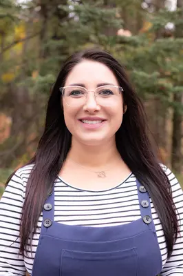 Smiling person outdoors in front of trees, wearing striped shirt and overalls.