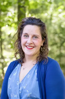 Smiling person with curly hair in a blue cardigan, forest background.