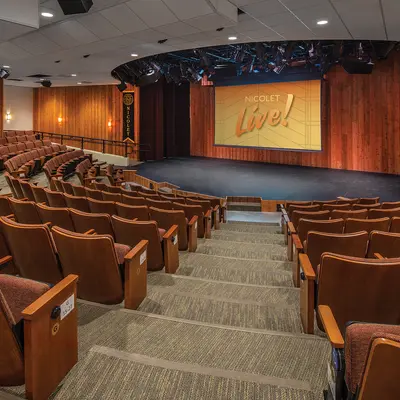 Theatre with rows of brown seats facing a stage and projector screen reading "Nicolet Live."