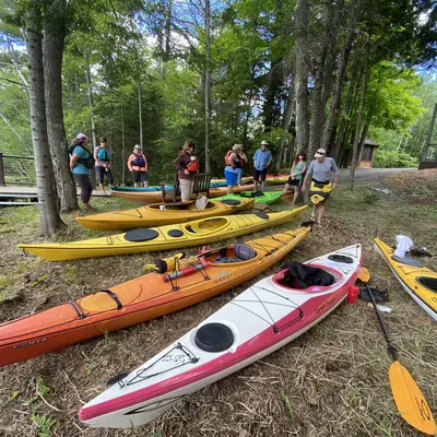 Colorful kayaks on grass, surrounded by people near trees and a wooden cabin.