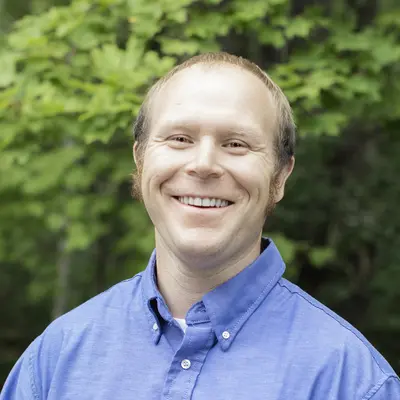 Smiling man in a blue shirt outdoors with lush greenery background.