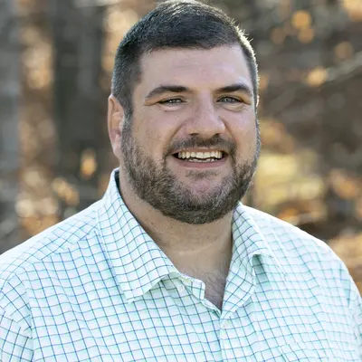 Smiling man in a checked shirt outdoors, trees in the background.