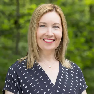Woman smiling in a forest setting, wearing a navy patterned blouse.