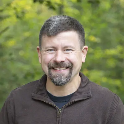 Smiling man outdoors with green leafy background.