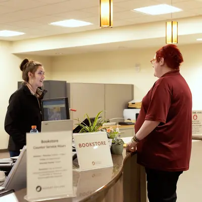 Student and campus Bookstore employee talking at a curved desk in a well-lit office.