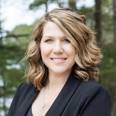 Smiling woman with wavy hair wearing a black blazer outdoors.