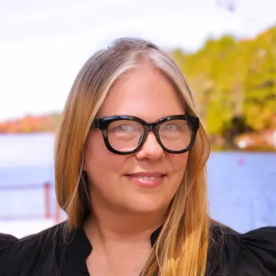 Smiling woman with glasses and long hair, standing outdoors by water.