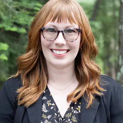 Smiling woman with red hair and glasses in a forest setting.
