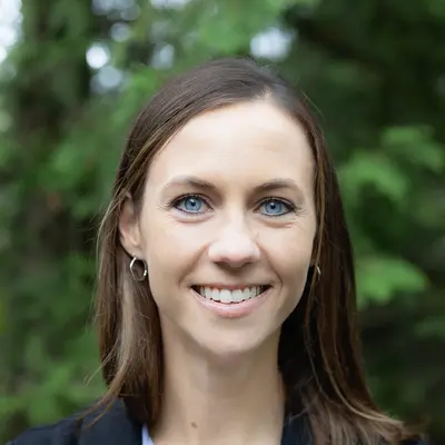 Smiling woman with brown hair, wearing a black blazer, outdoors.
