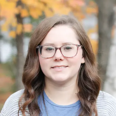 Woman with glasses smiling, wearing a blue shirt and cardigan, autumn trees in the background.