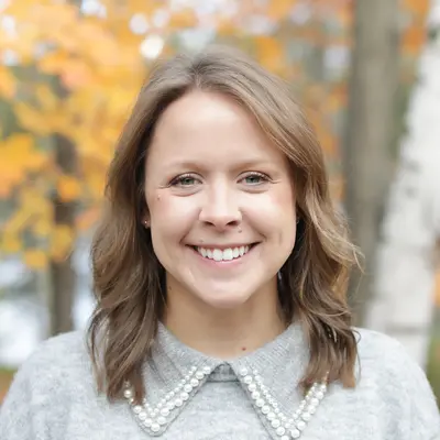 Smiling woman in a gray sweater outdoors with fall foliage in the background.