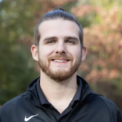 Smiling man with a topknot, wearing a black jacket, outdoors.