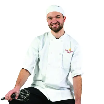 Chef in a white coat and hat sitting on a stool, holding a whisk.