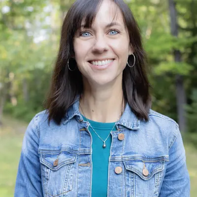 Smiling woman in a denim jacket outdoors with green trees.