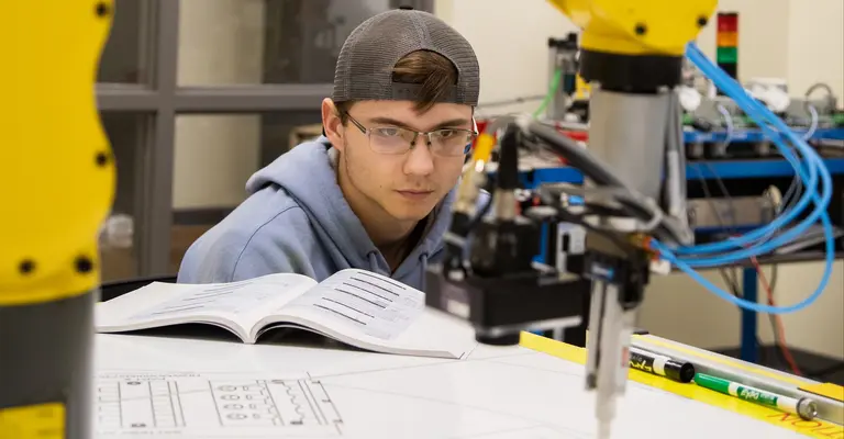 Student observes a robotic arm drawing on paper in a lab setting.