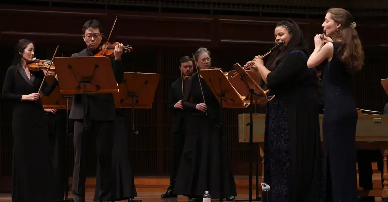 Musicians perform on stage, playing violins and flutes, wearing formal black attire.