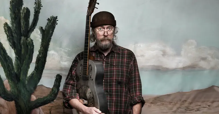 Charlie Parr, man in a plaid shirt holding a guitar, standing in a desert-themed setting with cacti.