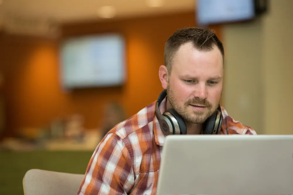 Male student working on laptop, wearing headphones and a plaid shirt.