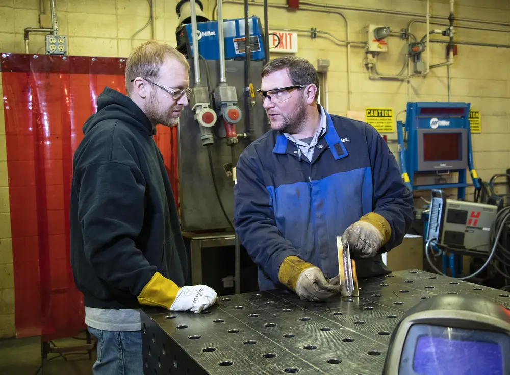 Welding student and instructor discussing metal work in a workshop near a metal table.