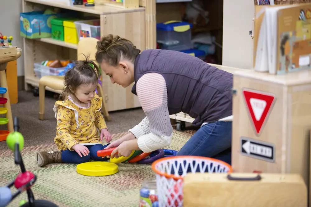 Early Childhood Education student and toddler playing with toys in a colorful classroom.