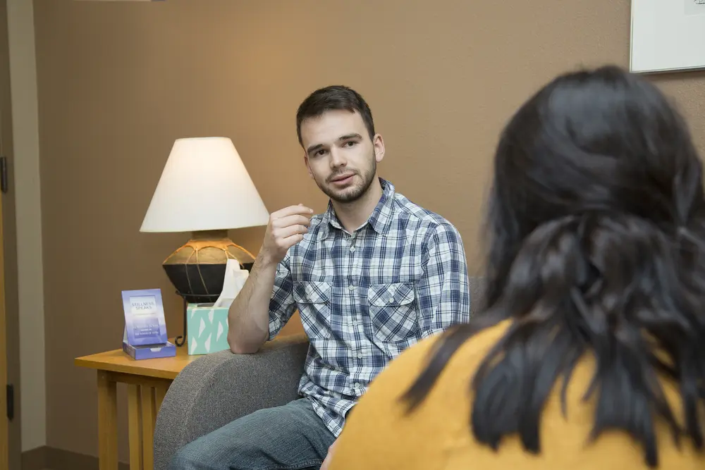 Man in plaid shirt speaking, seated in a cozy room with a lamp and tissue box.