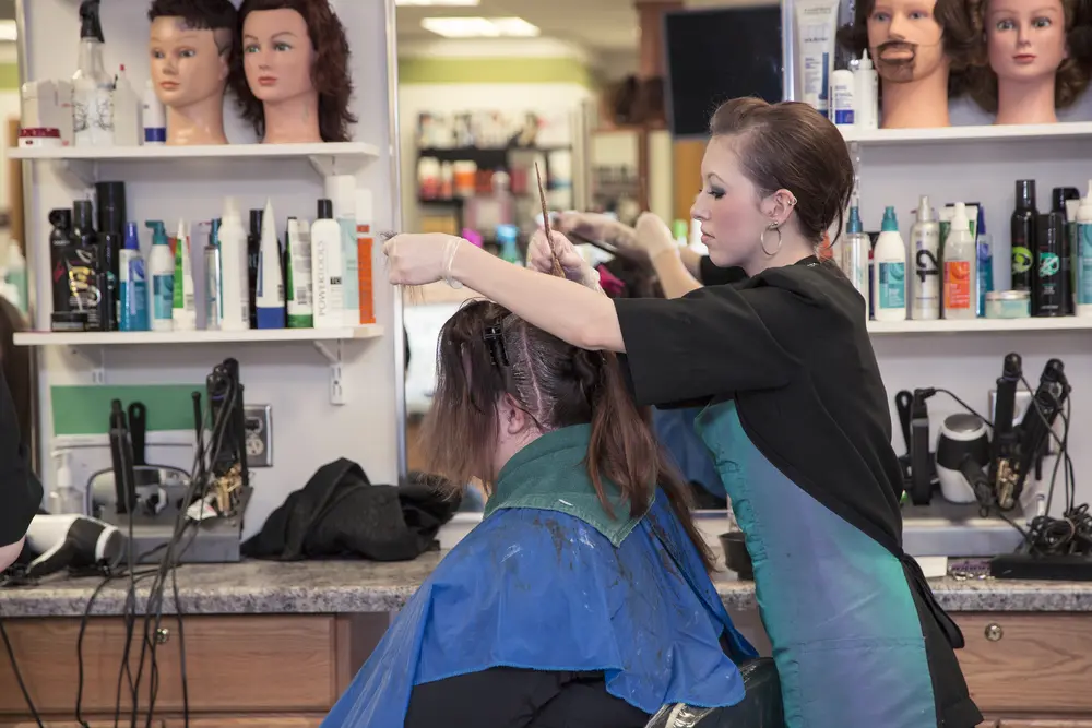 Female Cosmetology student dying a female client's hair in a salon with shelves of products.