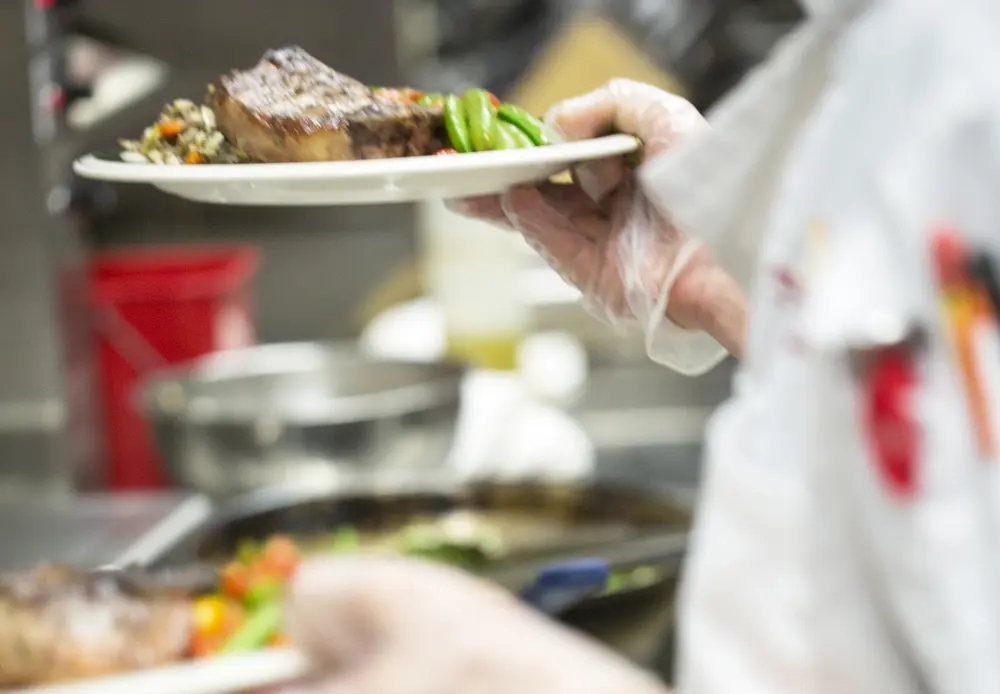 Culinary student holding plates of food in a busy kitchen.