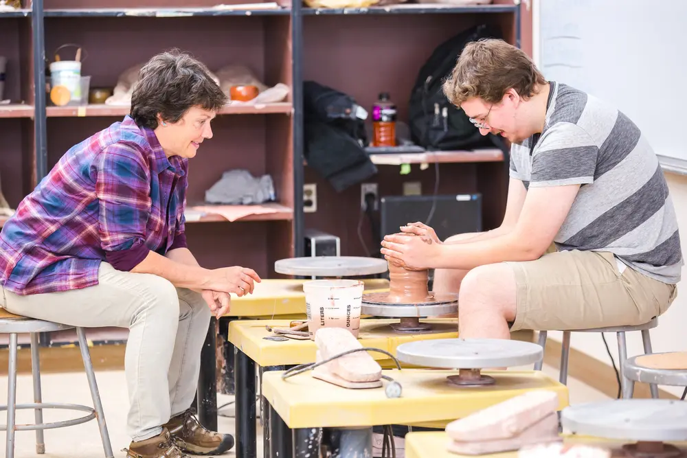 Two people sculpting clay on pottery wheels in a bright classroom.