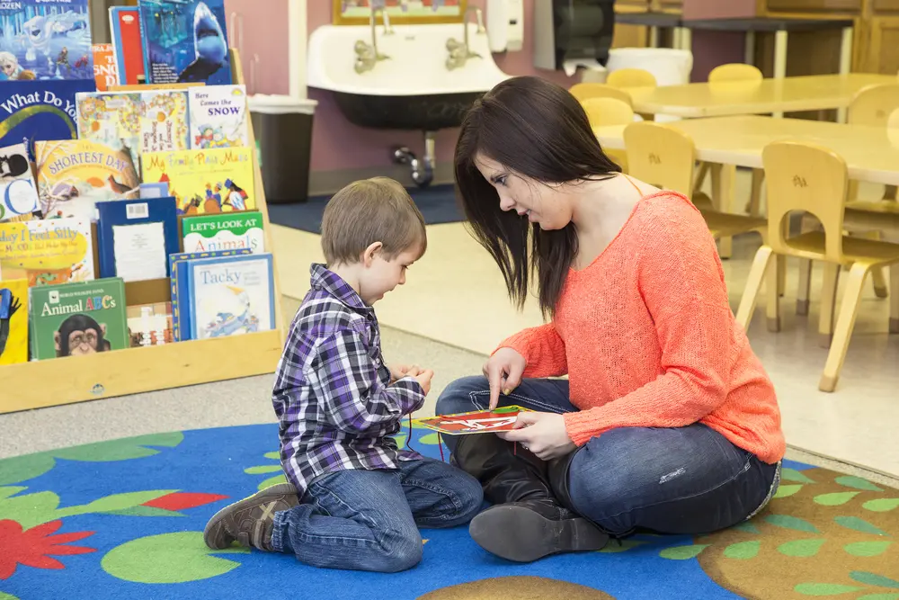 Early Childhood Education student and child reading a book on a colorful classroom rug.