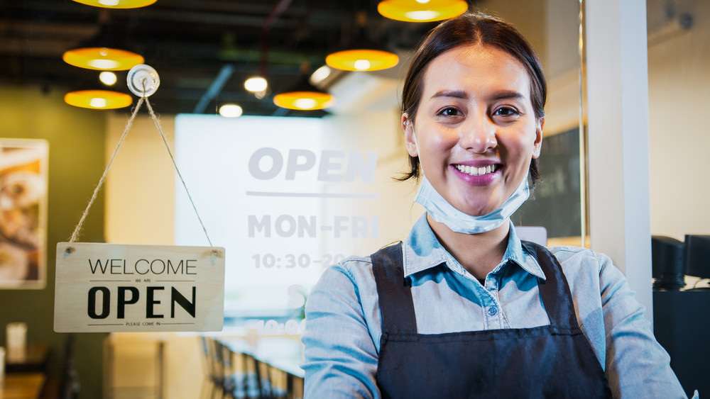 small business owner in front of open sign smiling with mask off of face