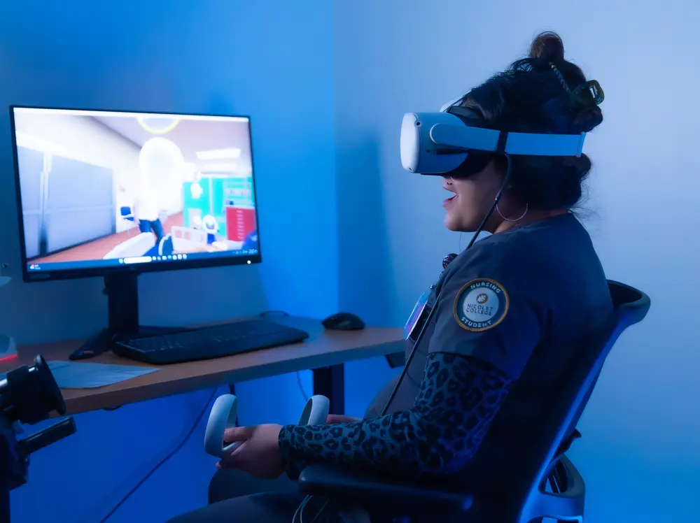 Nursing student using VR headset and controllers, seated by a computer monitor.