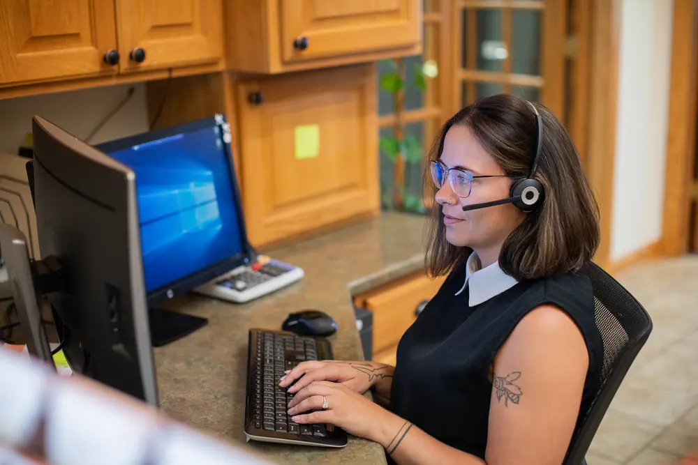 A woman with a headset works at a computer in an office setting.