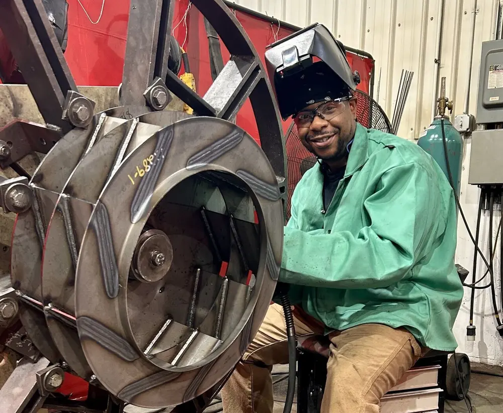 Man in green welding jacket, smiling near large metal machinery indoors.